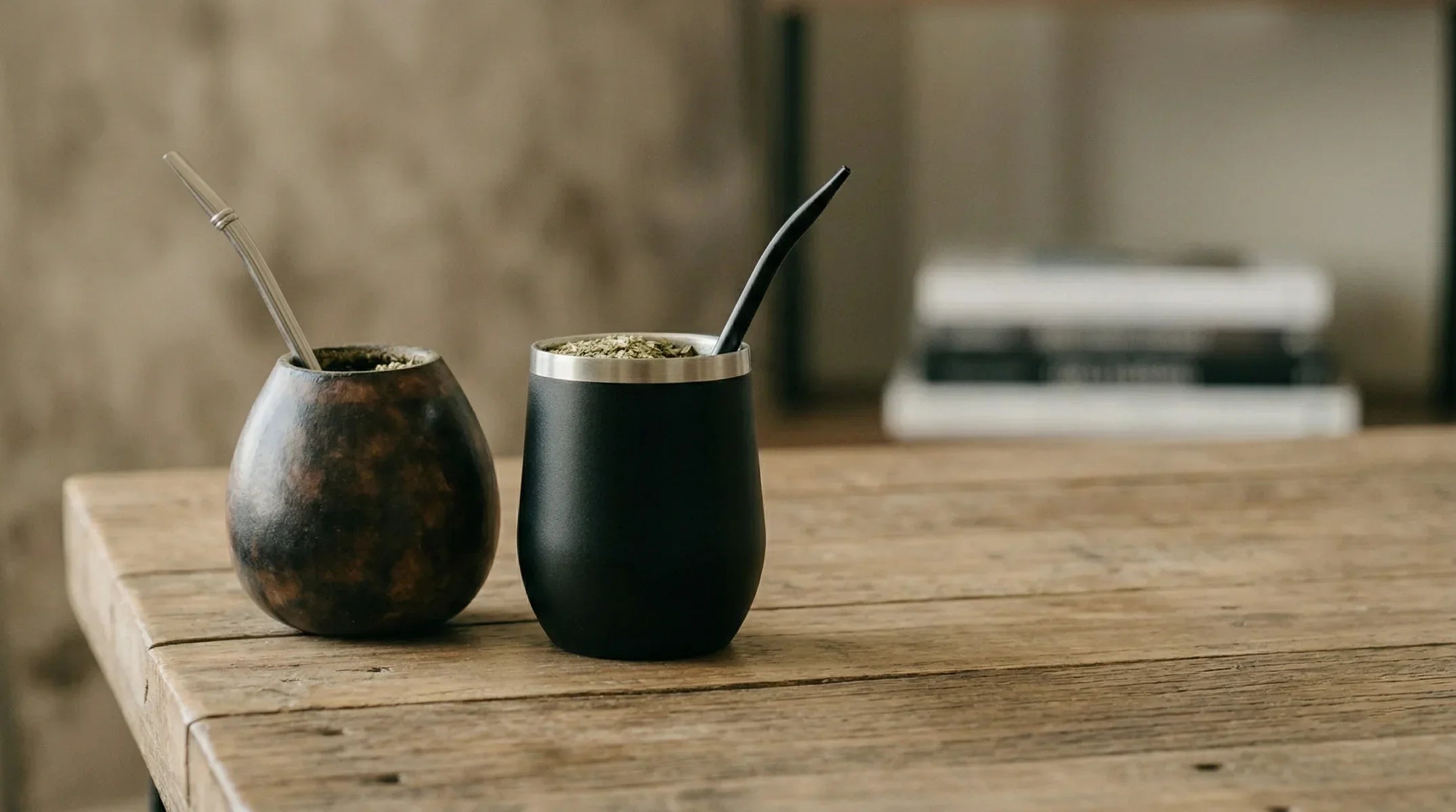 A side-by-side comparison of two yerba mate cups on a rustic wooden table. On the left a traditional dark porous calabaza gourd with a silver bombilla. On the right a modern matte black stainless steel mate cup with a black bombilla