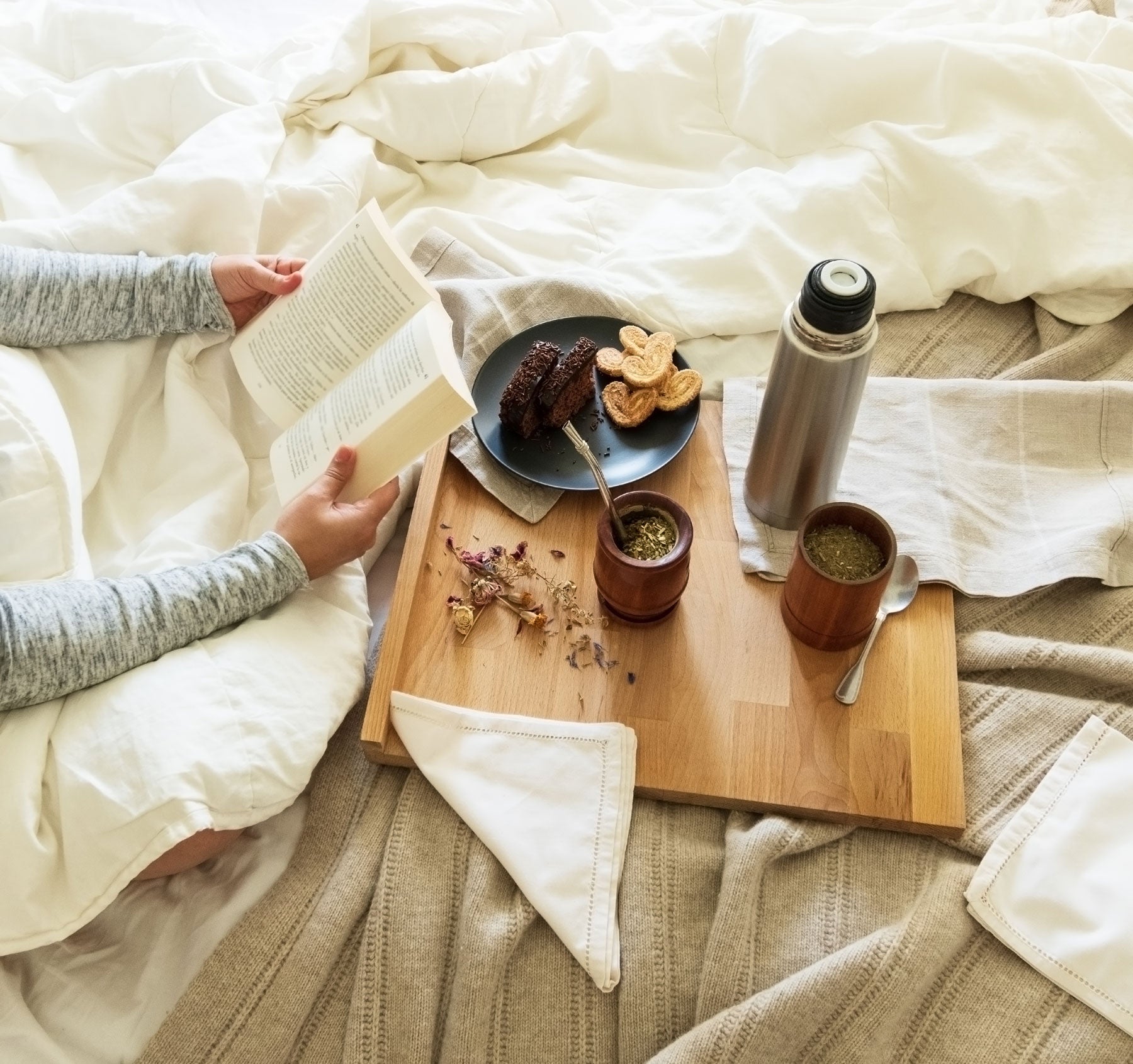 Person reading a book on a bed with a wooden tray holding a mate set and some pastries.