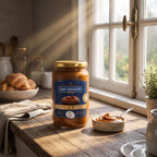 A sunlit glass jar of San Ignacio Dulce de Leche Traditional (450g) on a wooden kitchen counter, next to a small bowl of sauce with a spoon. A croissant and a multi-paned window are in the background.