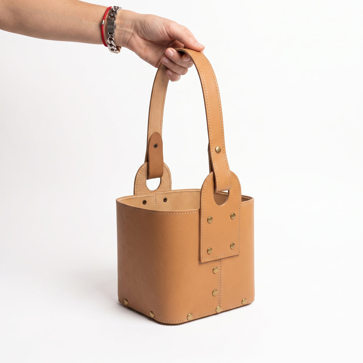 A hand holding a tan leather Criolla Matera by its top handle, featuring reinforced brass rivets along the side straps and base, set against a clean white background.
