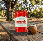 A red and white bag of Amanda Verba Mate sits on a rustic wooden picnic table outdoors, next to a traditional brown mate cup with a metal bombilla, with a blurred park background featuring trees.