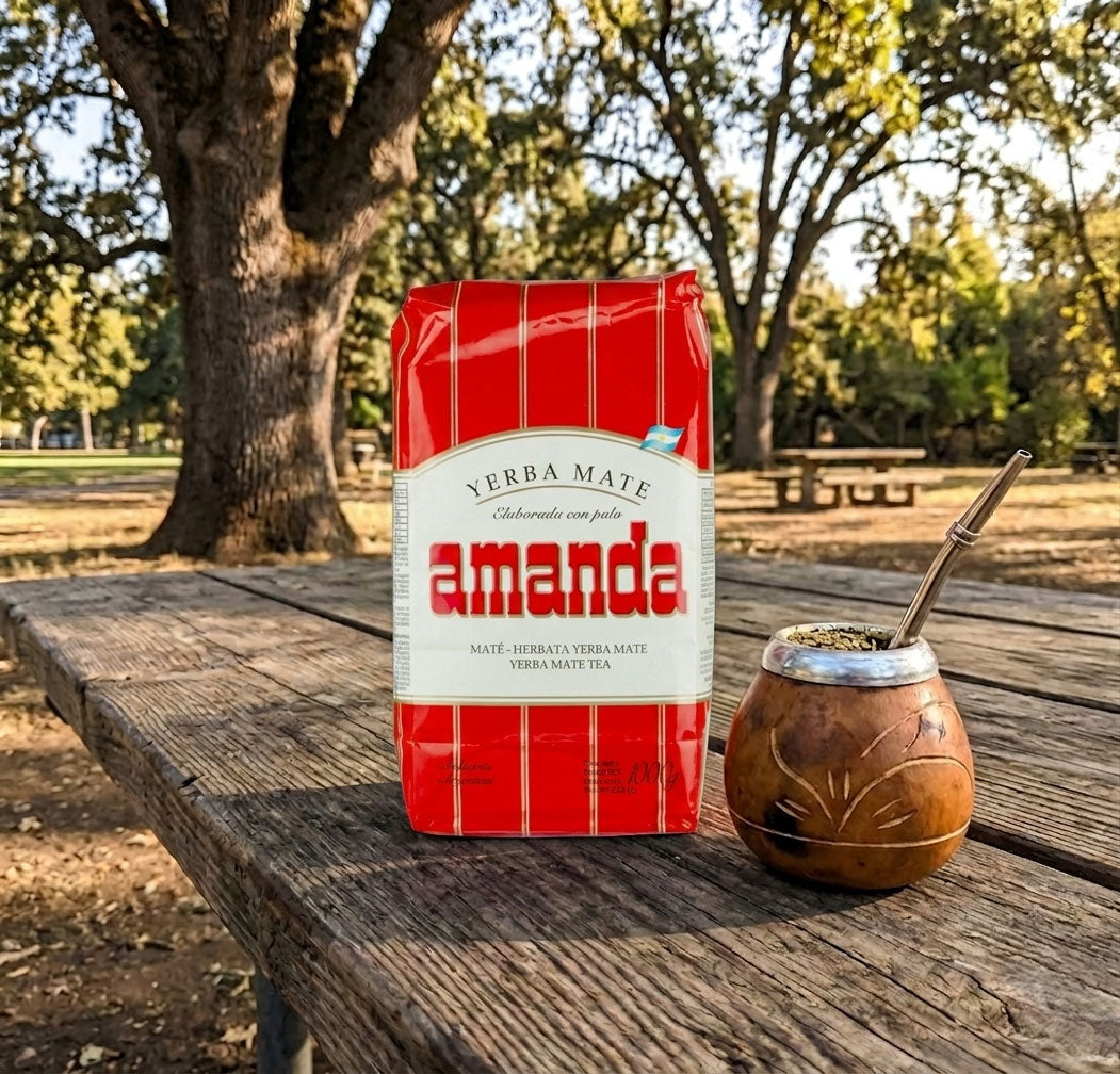 A red and white bag of Amanda Verba Mate sits on a rustic wooden picnic table outdoors, next to a traditional brown mate cup with a metal bombilla, with a blurred park background featuring trees.