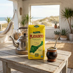 Playadito Yerba Mate 1kg bag on a wooden patio table alongside a traditional mate gourd with bombilla and a stainless steel kettle, with a sunlit ocean view in the background.