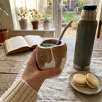 A hand holding the handcrafted carob wood Ranchera mate gourd, featuring a painted cowhide pattern and silver Southern Cross inlay, complete with a metal bombilla. The cozy indoor scene includes a green thermos, an open book, and alfajores on a rustic wooden table.