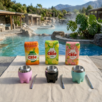 Three packages of CBSé flavoured yerba mate (Naranja, Limón, and Pomelo 500g) on a cloth next to three colourful mate gourds and bombillas, set outdoors by a pool with a mountain backdrop.