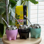 Colorful collection of modern no-cure Milonga mate gourds in Rose quartz, Obsidian, and Meadow with polished rims, displayed on a wooden stool next to Rosamonte Suave mild yerba mate.