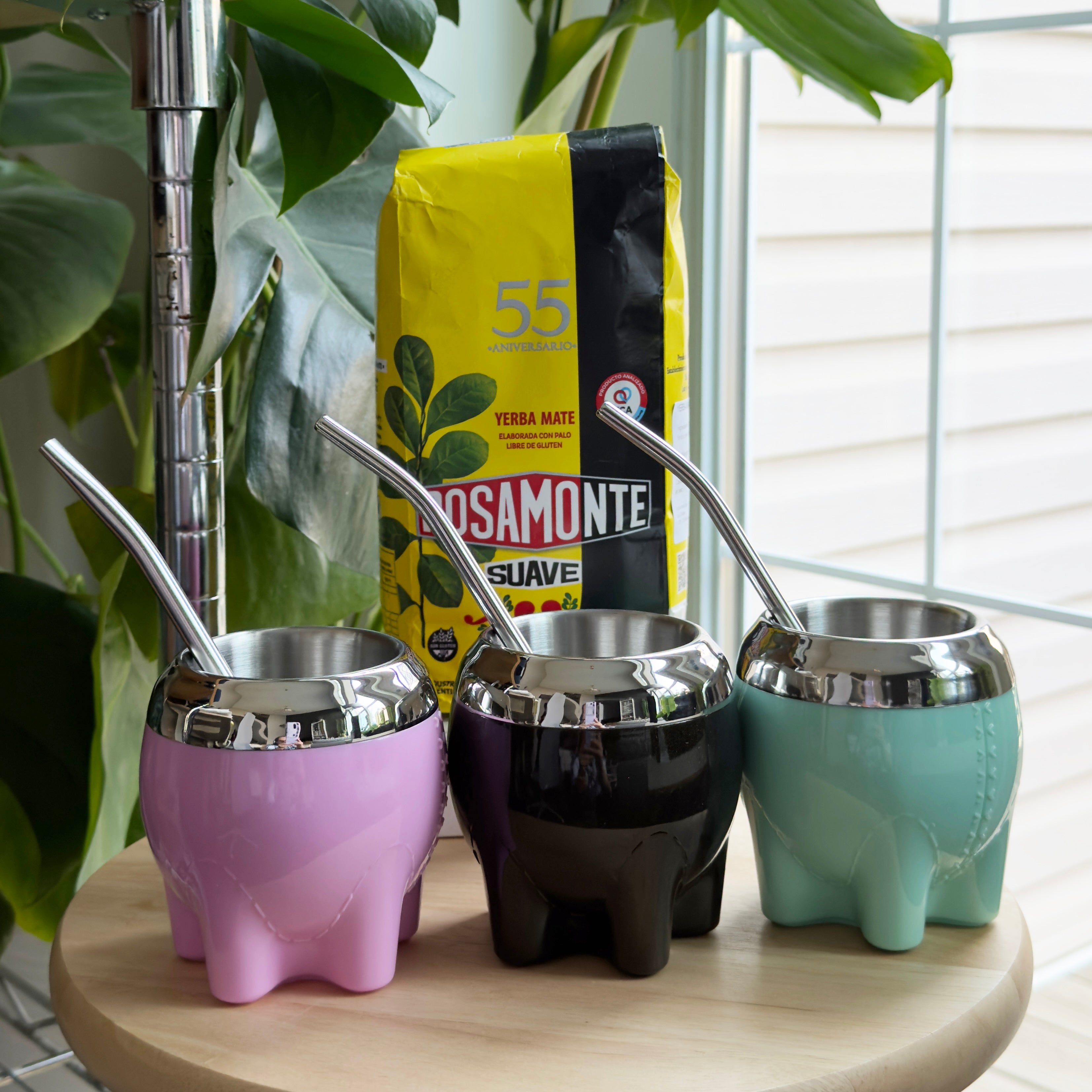Colorful collection of modern no-cure Milonga mate gourds in Rose quartz, Obsidian, and Meadow with polished rims, displayed on a wooden stool next to Rosamonte Suave mild yerba mate.