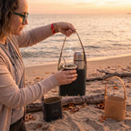 A woman on a beach holding a black leather Criolla Matera with a silver thermos and steaming mate gourd inside, while two additional leather materas in black and tan sit on the sand at sunset.