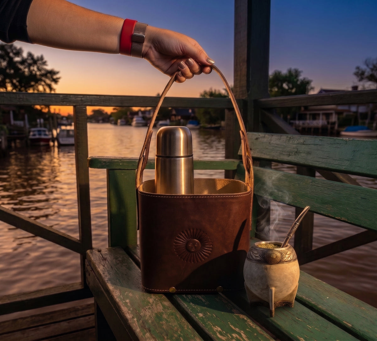 A hand holding the handle of a dark brown leather Criolla Matera on a wooden dock bench at sunset, containing a stainless steel thermos alongside a steaming mate gourd with a river background.