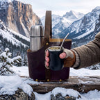 A dark brown leather Uruguaya Matera holding a stainless steel thermos on a snow-covered log, while a gloved hand holds a steaming black mate gourd with a silver bombilla straw against a scenic snowy mountain landscape.