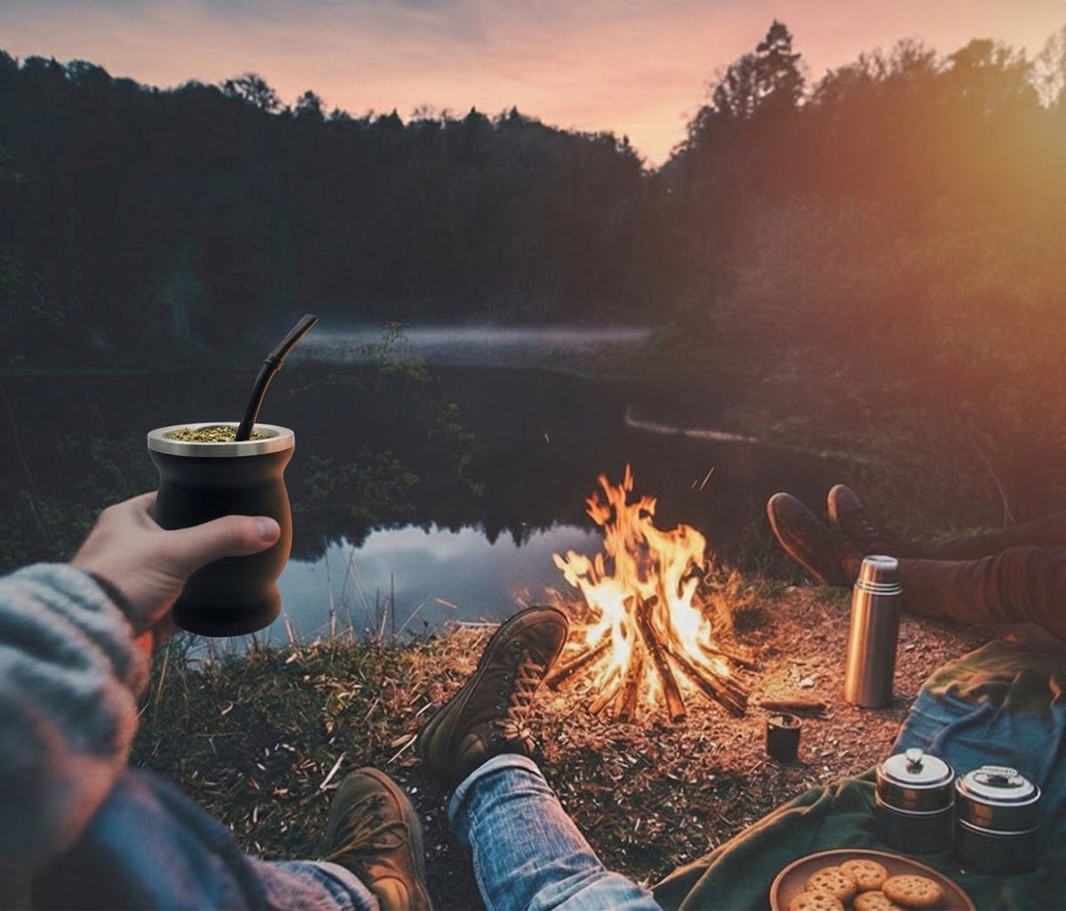 A cozy outdoor campfire scene at dusk by a lake, with a person holding a mate cup in the foreground and another person's legs visible near the fire. A thermos, mugs, and cookies sit on a blanket, with trees and misty water in the background.
