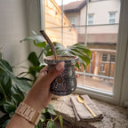 A hand holding the hand-chiseled Huella Artisan mate gourd filled with yerba mate and a metal bombilla. The scene is set by a bright window with a green plant and a gold spoon on the sill.