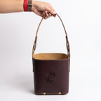 A hand holding a dark brown leather Criolla Matera by its top handle, showing the tan interior lining, brass rivets at the base, and an embossed Sun of May detail, against a plain white background.