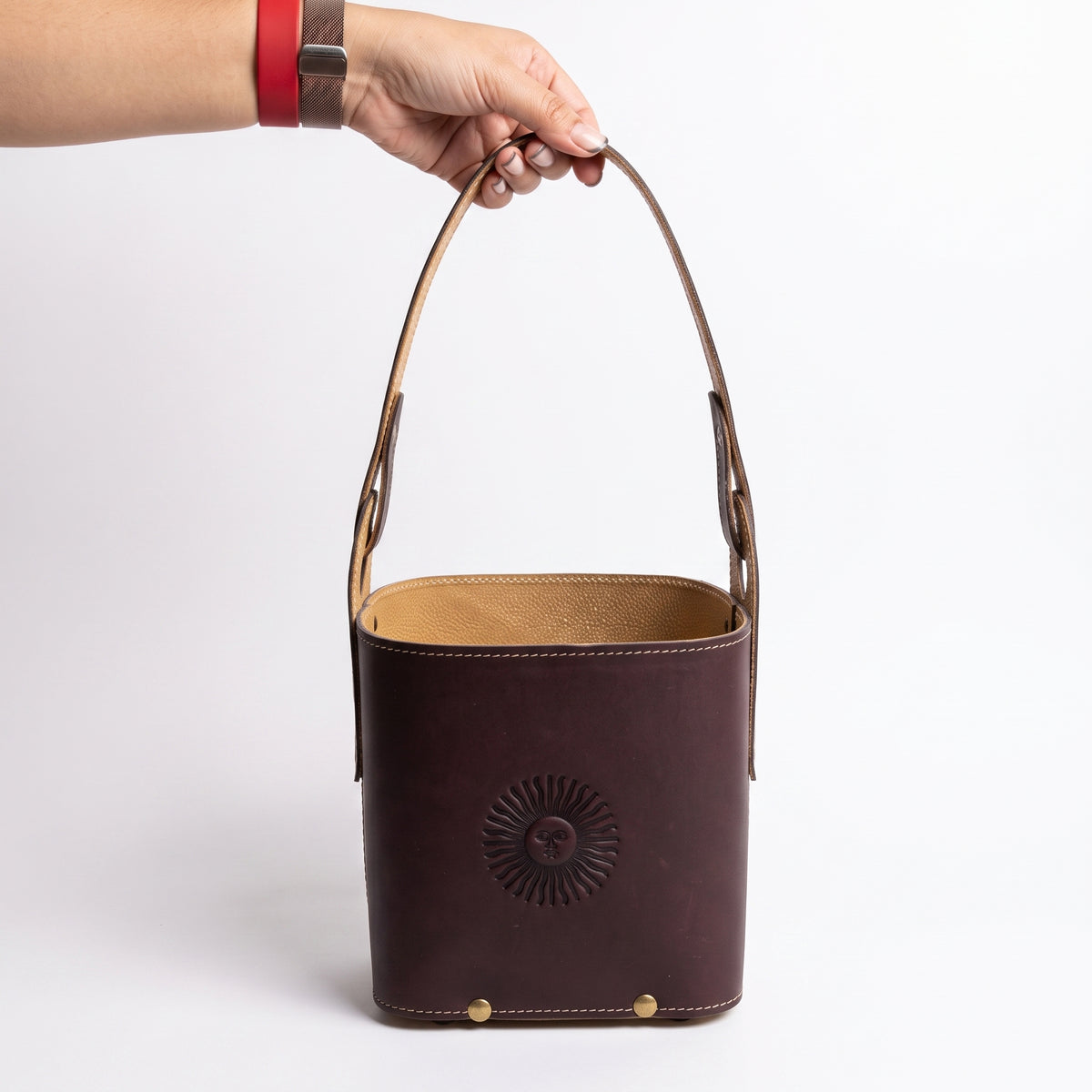 A hand holding a dark brown leather Criolla Matera by its top handle, showing the tan interior lining, brass rivets at the base, and an embossed Sun of May detail, against a plain white background.