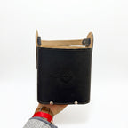 A hand holding the black leather Criolla Matera from the bottom, showing its arched top handle, prominent tan interior lining, three silver-tone studs on the front lower edge, and an embossed Sun of May emblem, set against a plain white background.