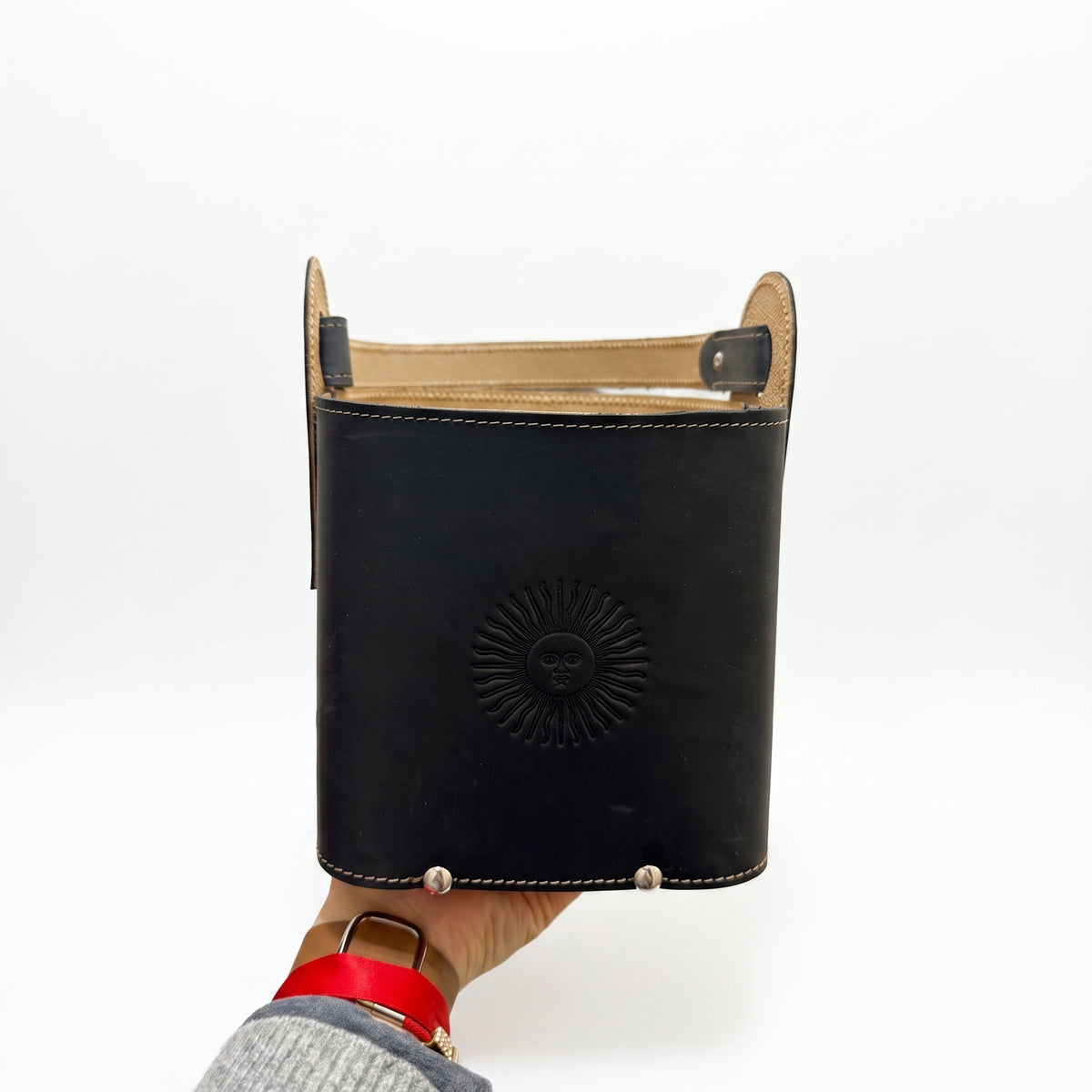 A hand holding the black leather Criolla Matera from the bottom, showing its arched top handle, prominent tan interior lining, three silver-tone studs on the front lower edge, and an embossed Sun of May emblem, set against a plain white background.