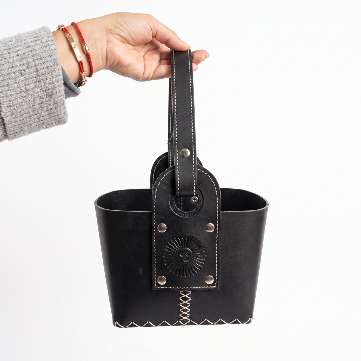 A hand holding a black leather Patria Matera by its top strap, featuring white contrast stitching, silver-tone studs, and an embossed Sun of May detail on the front, against a plain white background.