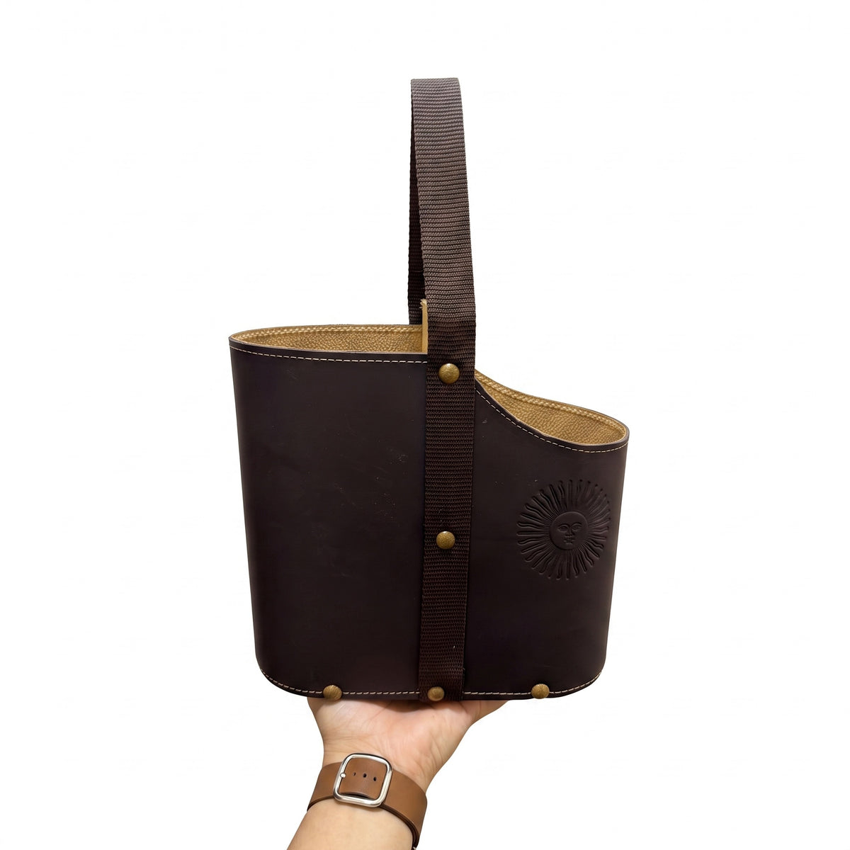 A dark brown leather Uruguaya Matera held from below by a hand, featuring a wide brown woven strap, brass-tone rivets, a tan leather lining, and an embossed Sun of May on the front, against a plain white background.