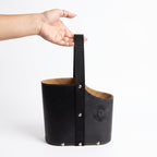 A hand holding a black leather Uruguaya Matera by its durable black woven webbing handle, showing the textured tan lining, silver metal rivets, and embossed Sun of May detail, set against a plain white background.