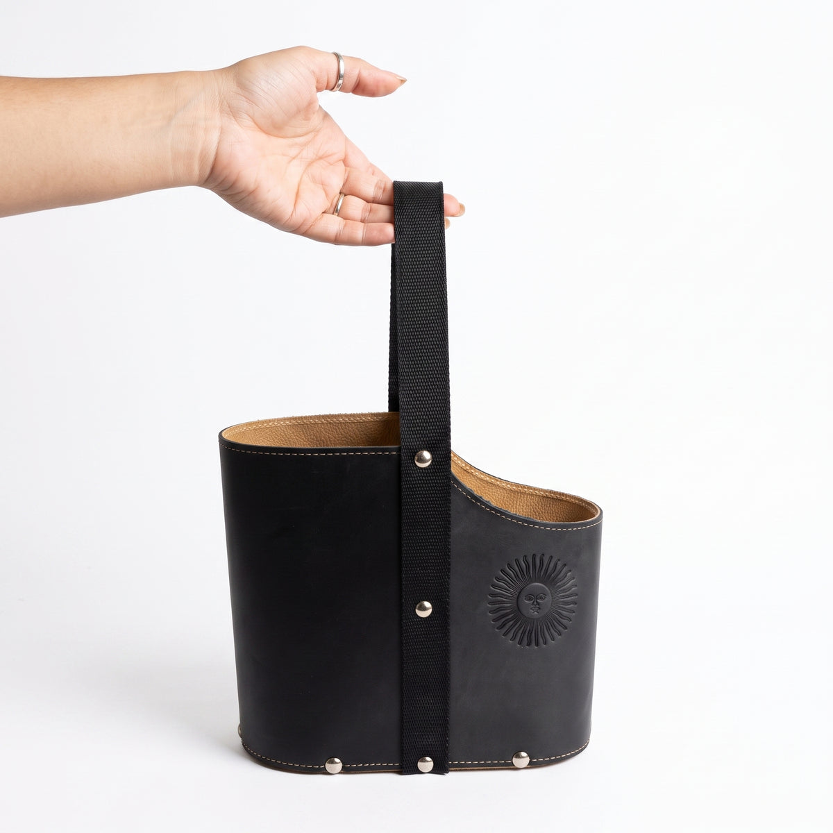 A hand holding a black leather Uruguaya Matera by its durable black woven webbing handle, showing the textured tan lining, silver metal rivets, and embossed Sun of May detail, set against a plain white background.