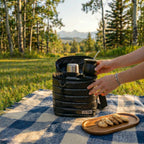 An outdoor lifestyle image showing 'The Nomad Carrier | Everyday Puffer Mate Bag', an open black quilted puffer-style insulated carrier bag, resting on a blue and white checkered picnic blanket. Inside the carrier is a stainless steel thermos, and a person's hands are holding a black mate gourd with a silver bombilla straw next to it. To the right is a small wooden tray with cookies. The scene is set in a grassy field with a gently blurred background of trees and mountains.