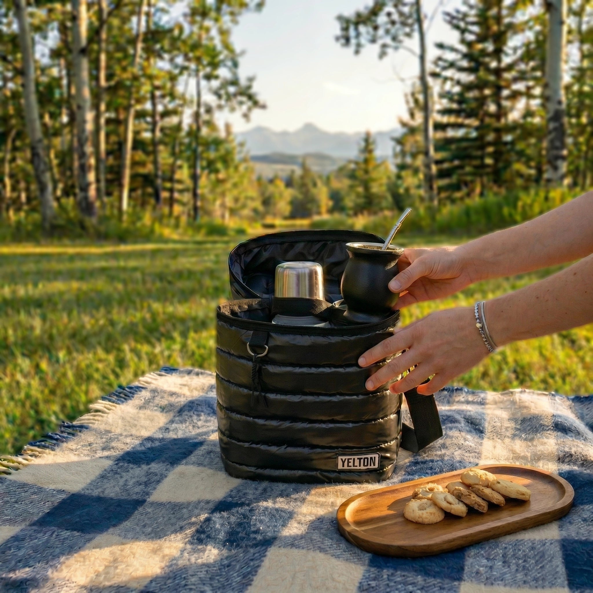 An outdoor lifestyle image showing 'The Nomad Carrier | Everyday Puffer Mate Bag', an open black quilted puffer-style insulated carrier bag, resting on a blue and white checkered picnic blanket. Inside the carrier is a stainless steel thermos, and a person's hands are holding a black mate gourd with a silver bombilla straw next to it. To the right is a small wooden tray with cookies. The scene is set in a grassy field with a gently blurred background of trees and mountains.