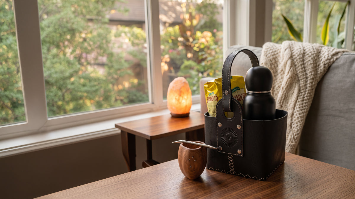 Black leather mate carrier bag with wooden mate cup, bombilla, thermos, and yerba mate bags on a wooden table.